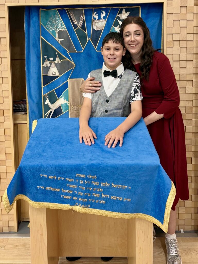 Young person and mother standing beside a Torah reading table in a synagogue, with a decorative blue ark curtain in the background ready for bar mitzvah.